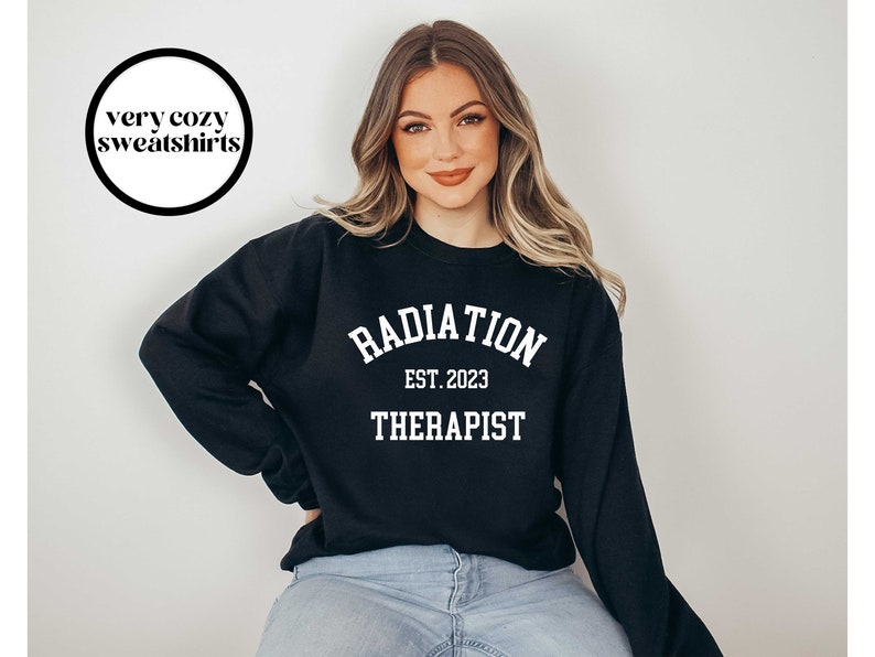 Puede incluir: Sudadera negra con texto blanco que dice "RADIATION EST. 2023 THERAPIST". Una mujer lleva la sudadera y unos vaqueros azules.