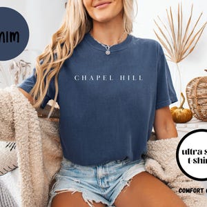 May include: A navy blue t-shirt with the words "Chapel Hill" printed in white. The shirt is part of the "Comfort Colors" range and is labelled "ultra soft t-shirts". A navy blue circle is in the upper left corner.