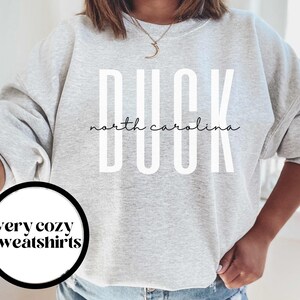 以下が含まれることがあります： グレーのトレーナーに「DUCK north carolina」という白い文字が書かれています。