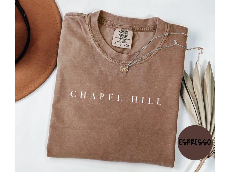 May include: A brown t-shirt with the words "CHAPEL HILL" in white block letters. A silver necklace with a small pendant rests on the shirt. A brown hat is visible in the upper left corner. The shirt is labeled "Espresso".