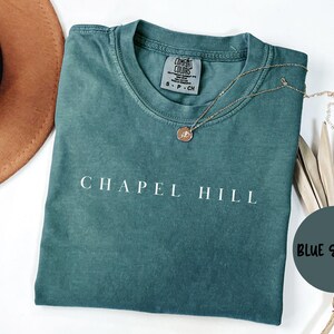 May include: Teal-coloured t-shirt with the text "CHAPEL HILL" in white. A gold necklace with a pendant rests on the shirt. A brown hat is visible in the background.
