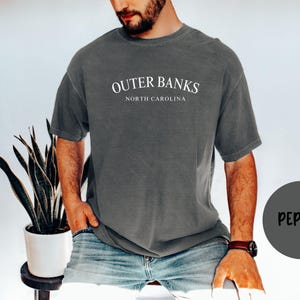 Puede incluir: Una camiseta gris con el texto "OUTER BANKS NORTH CAROLINA" en blanco. La camiseta es de color liso y tiene un corte relajado. La palabra "PEPPER" est&aacute; en un c&iacute;rculo gris en la esquina inferior derecha.