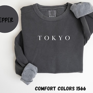May include: A dark gray sweatshirt with the word "TOKYO" in white letters. The sleeves are cuffed with a soft, gray material. A dark gray circle with the word "PEPPER" is in the upper left corner. The text "COMFORT COLORS 1566" is at the bottom.