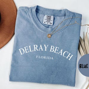 May include: A blue jean t-shirt with the text "Delray Beach, Florida" in white. The shirt has a distressed look and is made of a soft, comfortable fabric.