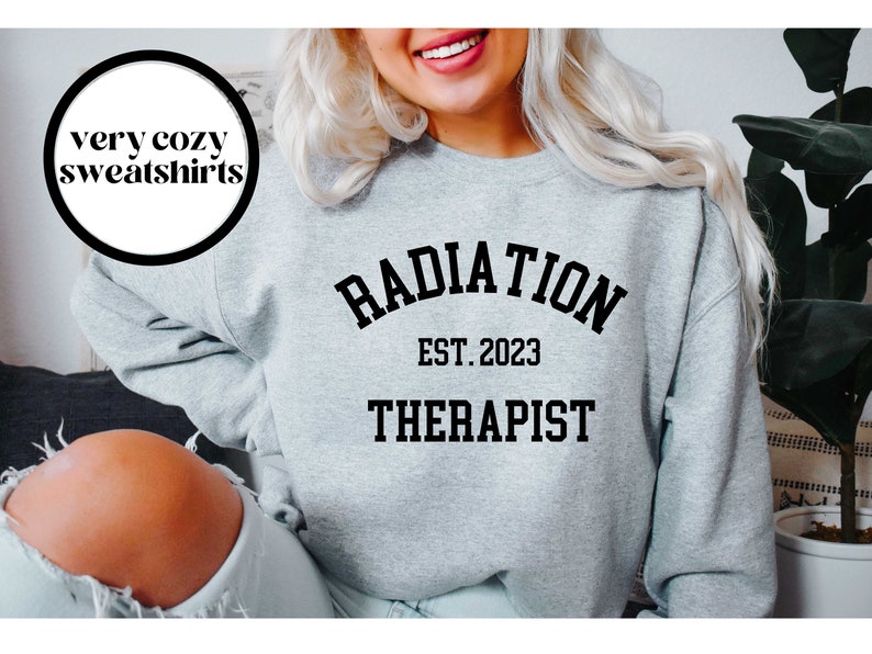 Puede incluir: Una sudadera gris con el texto "RADIATION THERAPIST EST. 2023" impreso en la parte delantera.