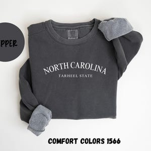 May include: A dark gray sweatshirt with the text "NORTH CAROLINA TARHEEL STATE" in white. The sweatshirt has a crew neck and the sleeves are cuffed. The word "PEPPER" is in a dark gray circle, and "COMFORT COLORS 1566" is at the bottom.