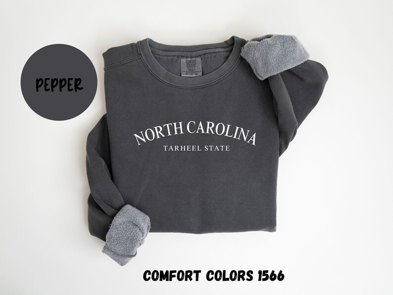May include: A dark gray sweatshirt with the text "NORTH CAROLINA TARHEEL STATE" in white. The sweatshirt has a crew neck and the sleeves are cuffed. The word "PEPPER" is in a dark gray circle, and "COMFORT COLORS 1566" is at the bottom.