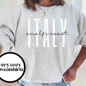 Puede incluir: Sudadera gris con el texto "ITALY amalfi coast" en letras blancas. La sudadera es de corte holgado y tiene cuello redondo.