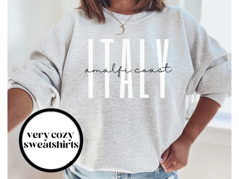 Puede incluir: Sudadera gris con el texto "ITALY amalfi coast" en letras blancas. La sudadera es de corte holgado y tiene cuello redondo.