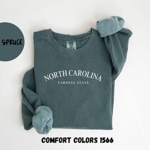 May include: Blue spruce sweatshirt with the text "NORTH CAROLINA TARHEEL STATE" in white. The sweatshirt has rolled sleeves and the words "COMFORT COLORS 1566" at the bottom. A blue circle with the words "BLUE SPRUCE" is in the upper left.