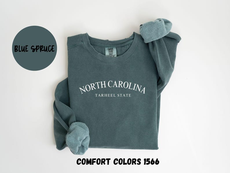 May include: Blue spruce sweatshirt with the text "NORTH CAROLINA TARHEEL STATE" in white. The sweatshirt has rolled sleeves and the words "COMFORT COLORS 1566" at the bottom. A blue circle with the words "BLUE SPRUCE" is in the upper left.
