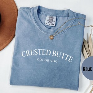 May include: A blue jean colored t-shirt with the text "CRESTED BUTTE COLORADO" in white. A gold necklace with a pendant is on the shirt. A brown hat and dried plants are in the background.