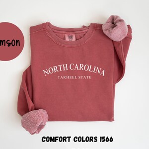May include: Crimson-colored sweatshirt with the text "NORTH CAROLINA TARHEEL STATE" in white. The sweatshirt has a crew neck and long sleeves with a soft, fuzzy cuff. The words "COMFORT COLORS 1566" are at the bottom.