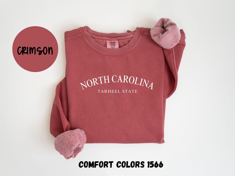 May include: Crimson-colored sweatshirt with the text "NORTH CAROLINA TARHEEL STATE" in white. The sweatshirt has a crew neck and long sleeves with a soft, fuzzy cuff. The words "COMFORT COLORS 1566" are at the bottom.