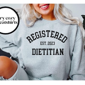 May include: A light gray sweatshirt with the words "REGISTERED DIETITIAN EST. 2023" in black block letters. The sweatshirt also has a circular graphic that says "very cozy sweatshirts".