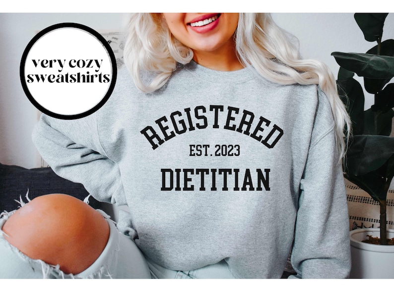 May include: A light gray sweatshirt with the words "REGISTERED DIETITIAN EST. 2023" in black block letters. The sweatshirt also has a circular graphic that says "very cozy sweatshirts".