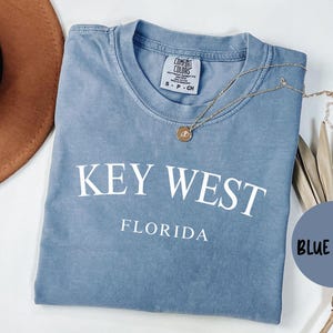 May include: A blue jean t-shirt with the text "KEY WEST FLORIDA" in white. The shirt is made of a soft, comfortable material and has a distressed look.