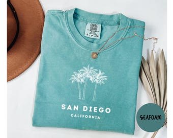 May include: Seafoam green t-shirt with a white palm tree graphic and the words "San Diego California". A gold necklace and a brown hat are next to the shirt. The shirt has a Comfort Colors tag.