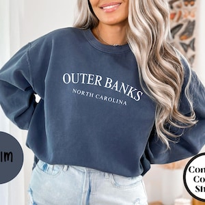 May include: Denim blue sweatshirt with the text "Outer Banks North Carolina" in white. The sweatshirt is part of the Comfort Colors Shirts collection. A denim-colored circle with the word "Denim" is in the lower left corner.