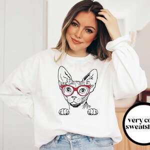 May include: White sweatshirt with a black and white illustration of a Sphynx cat wearing red glasses. The text "very cozy sweatshirts" is on a black circle in the bottom right corner of the image.