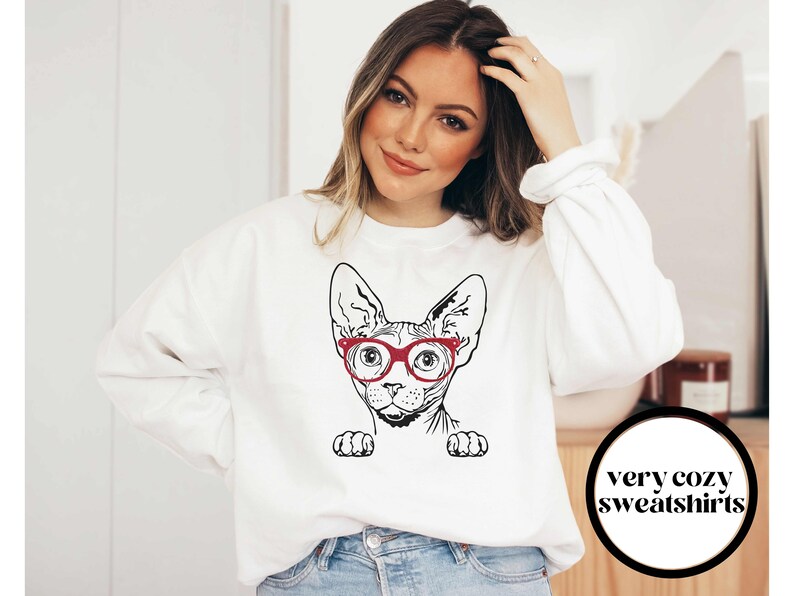 May include: White sweatshirt with a black and white illustration of a Sphynx cat wearing red glasses. The text "very cozy sweatshirts" is on a black circle in the bottom right corner of the image.