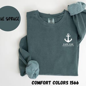 May include: A blue spruce Comfort Colors 1566 sweatshirt with a white anchor graphic and the text "Cape Cod Massachusetts." The sweatshirt has long sleeves and a crew neck. The color name "Blue Spruce" is in a circle.