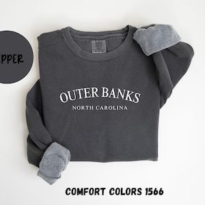 May include: A dark gray sweatshirt with the text "Outer Banks North Carolina" in white. The sweatshirt has a crew neck and long sleeves with gray fleece cuffs. A dark gray circle with the word "Pepper" is in the upper left corner. The text "Comfort Colors 1566" is at the bottom.
