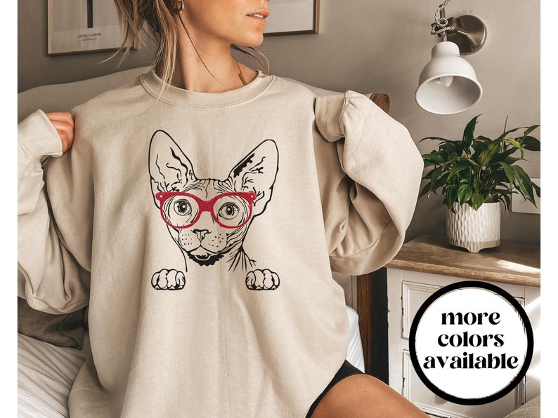 May include: A beige sweatshirt with a black and white illustration of a Sphynx cat wearing red glasses. The text "more colors available" is visible in the lower right corner of the image.