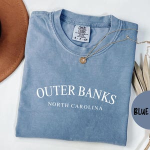 Puede incluir: Una camiseta azul vaquera con las palabras "Outer Banks North Carolina" impresas en blanco. Tambi&eacute;n se ven un collar dorado y un sombrero marr&oacute;n. La camiseta est&aacute; doblada y tiene una etiqueta Comfort Colors.