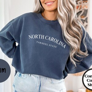May include: Denim blue sweatshirt with the text "NORTH CAROLINA TARHEEL STATE" in white. The sweatshirt is part of the "Comfort Colors Shirts" collection. The word "DENIM" is in a circle on the left.