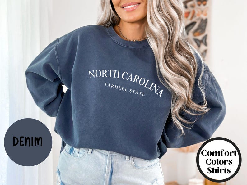 May include: Denim blue sweatshirt with the text "NORTH CAROLINA TARHEEL STATE" in white. The sweatshirt is part of the "Comfort Colors Shirts" collection. The word "DENIM" is in a circle on the left.