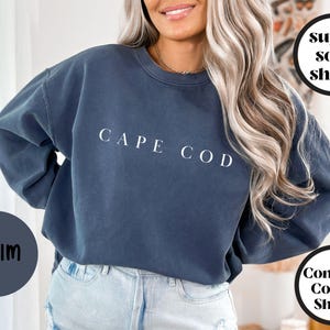 May include: A denim blue crew neck sweatshirt with the white text "CAPE COD". The image also includes the text "super soft shirts" and "Comfort Colors Shirts". The sweatshirt appears to be made of a soft material.