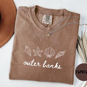 May include: Espresso-colored t-shirt with white seashell graphics and the words "outer banks." A silver necklace with a pendant rests on the shirt. A brown hat and dried plants are visible in the background.
