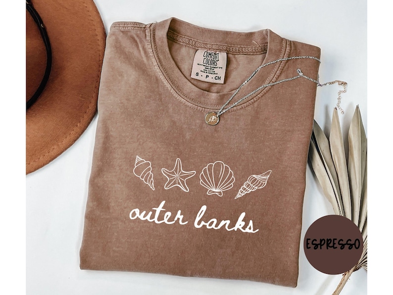 May include: Espresso-colored t-shirt with white seashell graphics and the words "outer banks." A silver necklace with a pendant rests on the shirt. A brown hat and dried plants are visible in the background.