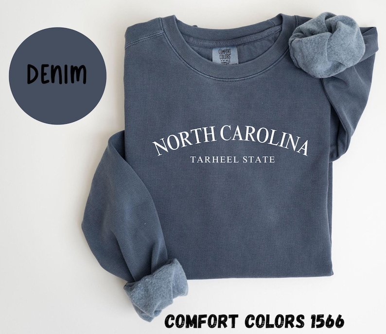 May include: A denim blue sweatshirt with the text "NORTH CAROLINA TARHEEL STATE" in white. The sweatshirt has a crew neck and long sleeves with cuffed ends. A denim blue circle with the word "DENIM" is in the upper left corner. The text "COMFORT COLORS 1566" is at the bottom.