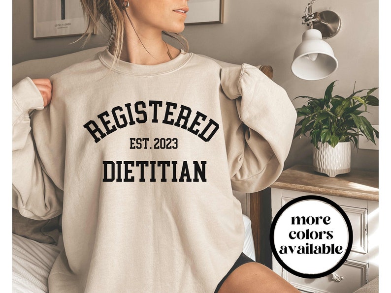 May include: Beige sweatshirt with the text "REGISTERED EST. 2023 DIETITIAN" in black, arched font. The sweatshirt has long sleeves and a crew neck. A circular graphic in the bottom right corner says "more colors available".