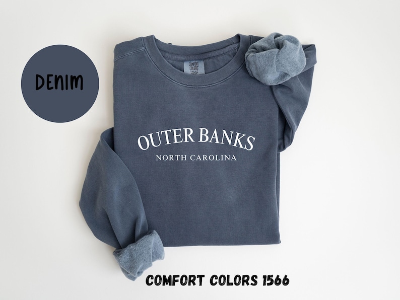 May include: Denim blue sweatshirt with the text "Outer Banks North Carolina" in white. The sweatshirt is a Comfort Colors 1566 style. The word "Denim" is in a dark blue circle.