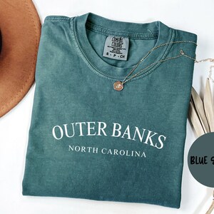 Puede incluir: Una camiseta azul abeto con las palabras "Outer Banks North Carolina" en blanco. La camiseta est&aacute; doblada y tiene un collar dorado encima. Un sombrero marr&oacute;n y hojas decorativas tambi&eacute;n est&aacute;n en la imagen.