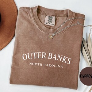Puede incluir: Camiseta color espresso con las palabras "Outer Banks North Carolina" en blanco. Un collar plateado con un colgante descansa sobre la camiseta. Un sombrero marr&oacute;n es visible en la esquina superior izquierda. La camiseta est&aacute; doblada.