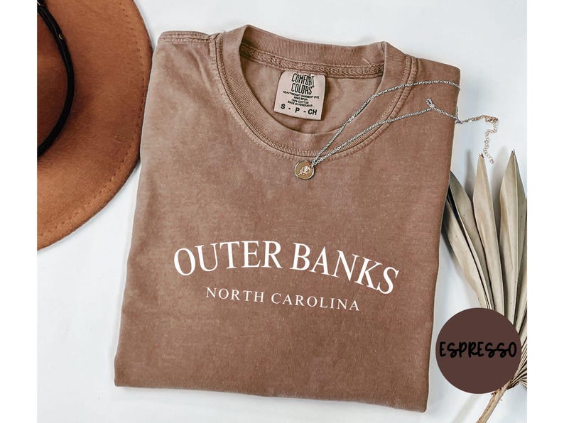 Puede incluir: Camiseta color espresso con las palabras "Outer Banks North Carolina" en blanco. Un collar plateado con un colgante descansa sobre la camiseta. Un sombrero marr&oacute;n es visible en la esquina superior izquierda. La camiseta est&aacute; doblada.