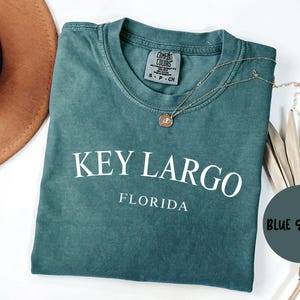 May include: A teal blue t-shirt with the text "KEY LARGO FLORIDA" in white. The shirt is made of a soft, comfortable material and has a vintage look.