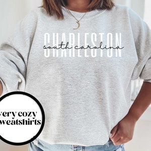 May include: A gray sweatshirt with the text "CHARLESTON south carolina" in white lettering. The sweatshirt is a relaxed fit and has a crew neck. The text "very cozy sweatshirts" is in a circle on the bottom left of the image.