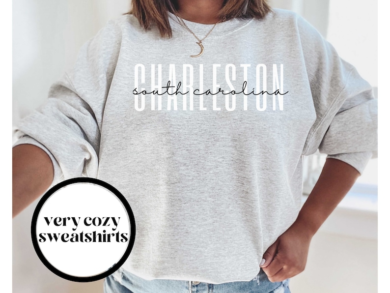 May include: A gray sweatshirt with the text "CHARLESTON south carolina" in white lettering. The sweatshirt is a relaxed fit and has a crew neck. The text "very cozy sweatshirts" is in a circle on the bottom left of the image.