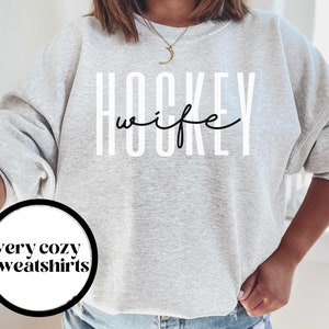 Op de afbeelding: Grijs sweatshirt met de tekst "HOCKEY wife" in witte letters met een zwarte omtrek. De tekst is in een modern, gestileerd lettertype.