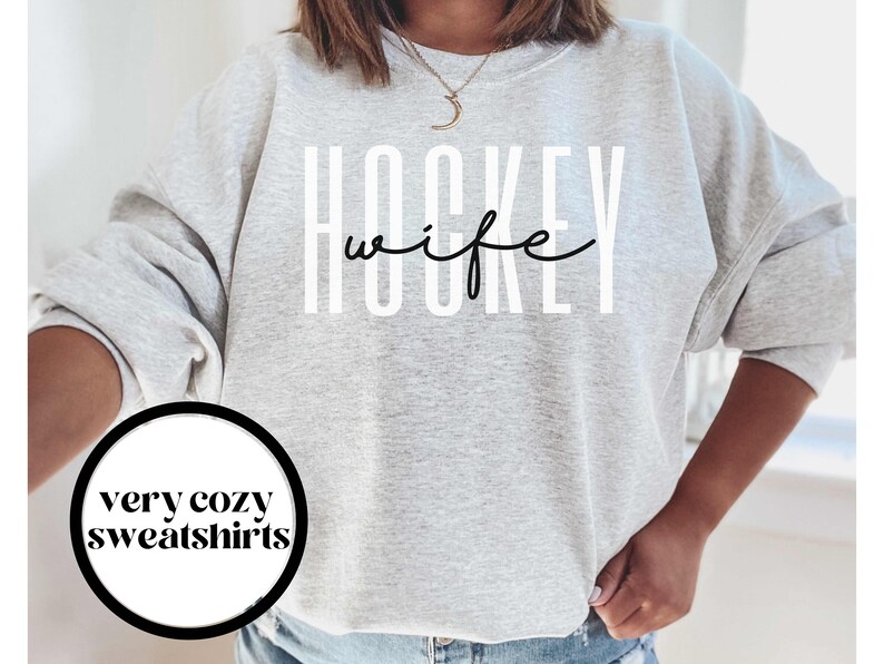 Op de afbeelding: Grijs sweatshirt met de tekst "HOCKEY wife" in witte letters met een zwarte omtrek. De tekst is in een modern, gestileerd lettertype.