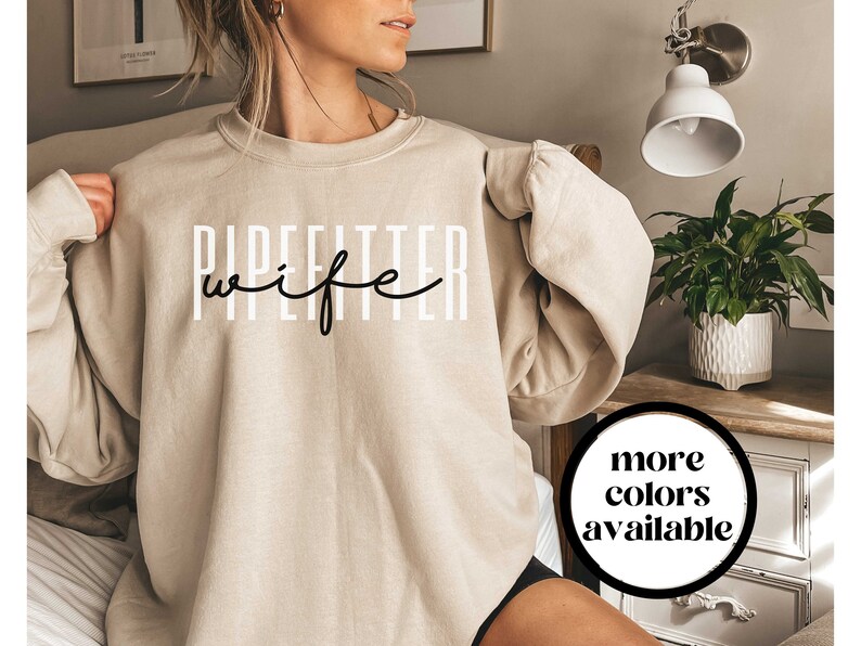 May include: A beige sweatshirt with the text "PIPEFITTER wife" in black lettering. The text is in a stylized font with the word "wife" in cursive. The sweatshirt is being worn by a person. The text "more colors available" is in a circle in the lower right corner of the image.