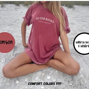 Puede incluir: Camiseta color carmes&iacute; con el texto "Outer Banks North Carolina" en blanco. La camiseta est&aacute; etiquetada como "ultra soft t shirts" y "Comfort Colors 1717". La modelo est&aacute; sentada sobre una superficie arenosa.