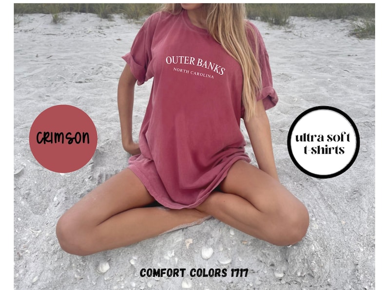 Puede incluir: Camiseta color carmes&iacute; con el texto "Outer Banks North Carolina" en blanco. La camiseta est&aacute; etiquetada como "ultra soft t shirts" y "Comfort Colors 1717". La modelo est&aacute; sentada sobre una superficie arenosa.