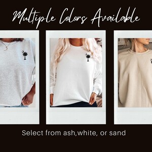 May include: Three photos showing a sweatshirt in three different colors: ash, white, and sand. Each sweatshirt features a small embroidered design of a palmetto tree with a crescent moon.