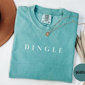 May include: Seafoam green t-shirt with the word "DINGLE" in white block letters. A gold necklace with a pendant rests on the shirt. A brown hat and a sprig of dried leaves are in the background.